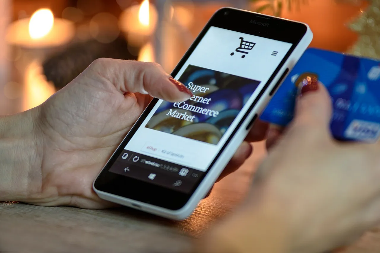 Ein Smartphone in der linken Hand einer Frau mit einem Onlineshop auf dem Display und in der rechten Hand eine unkenntlich gemachte Kreditkarte