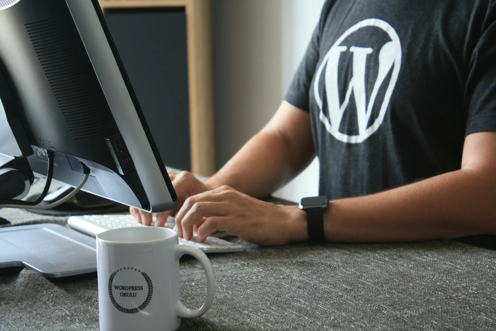 Ein Mann sitzt an einem Schreibtisch und tippt auf einer Computertastatur. Er trägt ein schwarzes T-Shirt mit dem WordPress-Logo. Auf dem Schreibtisch steht eine weiße Tasse mit der Aufschrift WordPress Entwickler